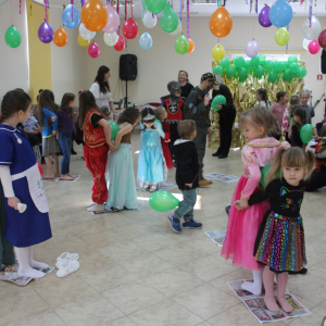 Pary dzieci biorą udział w zabawie zręcznościowej z zielonymi balonami w sali udekorowanej kolorowymi dekoracjami.