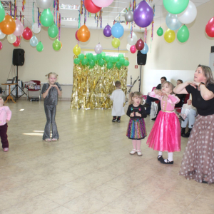 Grupa dzieci i opiekunka wykonują taneczne gesty w sali udekorowanej wiszącymi balonami.