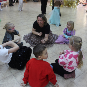 Grupa dzieci i siedząca z nimi na podłodze uśmiechnięta kobieta podczas wspólnej zabawy.