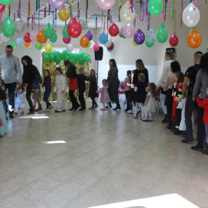 zieci i dorośli idą w korowodzie wzdłuż ścian udekorowanej sali, trzymając się za ręce lub ramiona.