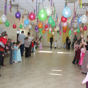 Dzieci i opiekunki stoją w dwóch rzędach wzdłuż ścian sali udekorowanej wiszącymi, kolorowymi balonami.