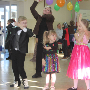 Dzieci i opiekunka wykonują taneczne gesty w rytm muzyki w udekorowanej sali.