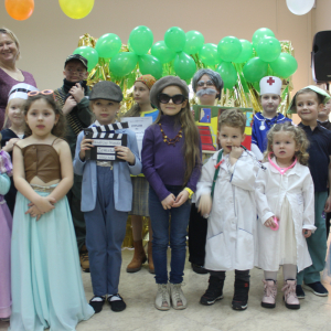 Grupa dzieci w przebraniach za różne zawody i postaci (m.in. lekarz, malarz, reżyser, pielęgniarka), stojąca na tle zielonych balonów i złotej kurtyny.