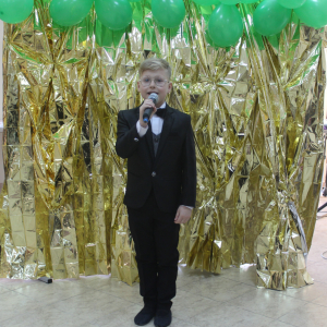 Chłopiec w garniturze z muchą trzymający mikrofon na tle złotej kurtyny.