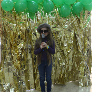 Dziewczynka w berecie i ciemnych okularach trzyma mikrofon na tle złotej dekoracji.