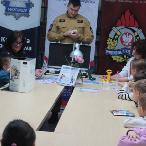 Spotkanie edukacyjne dzieci ze strażakiem i policjantką, z materiałami o bezpieczeństwie, w sali z banerami służb