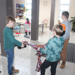 Trzej chłopcy w bibliotece rozciągają długi kawałek kolorowego materiału. W tle widać pozostałe dzieci pracujące przy stole oraz regały z książkami.