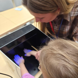Dorosła osoba pomaga dziecku włożyć dłonie do urządzenia z lampą UV podczas zajęć edukacyjnych.