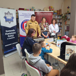Spotkanie edukacyjne dzieci ze strażakiem i policjantką oraz z panią z sanepidu, z materiałami o bezpieczeństwie, w sali z banerami służb