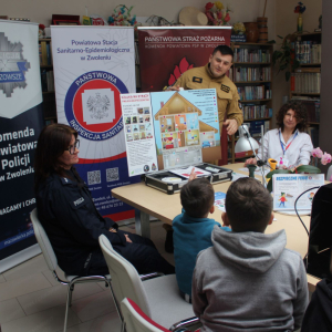 Strażak i policjantka oraz pani z sanepidu prowadzą zajęcia o bezpieczeństwie dla dzieci przy stole z materiałami edukacyjnymi.