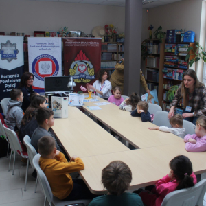 Strażak i policjantka oraz pani z sanepidu prowadzą zajęcia o bezpieczeństwie dla dzieci przy stole z materiałami edukacyjnymi.