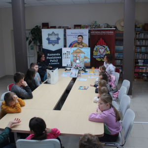 Spotkanie edukacyjne dzieci ze strażakiem i policjantką, z materiałami o bezpieczeństwie, w sali z banerami służb