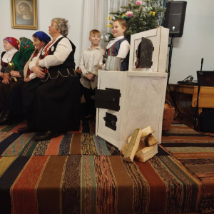 Dwójka dzieci stoi przy piecu. Kobieta w stroju ludowym siedzi na ławie