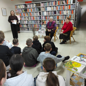Na zdjęciu wykonanym w czytelni widać grupę dzieci siedzących na dywanie. Przed nimi, na krzesłach, siedzą dwie panie. Obok nich stoi prowadząca, trzymająca w ręku kartkę z materiałami lub notatkami.