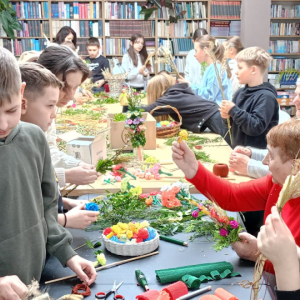 Dzieci stoją przy stole obok siedzą prowadzące zajęcia, wszyscy robią palmy wielkanocne.