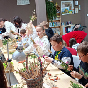 Przy stole siedzą dzieci wykonują palmy wielkanocne z materiałów które leżą przed nimi na stole.