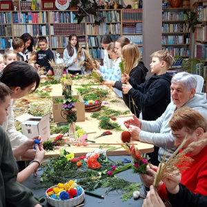 Przy stole siedzą uczestnicy zajęć wraz z prowadzącymi, niektórzy trzymają palmy ,przed nimi na stole leżą różne materiały m. in. zielone rośliny.