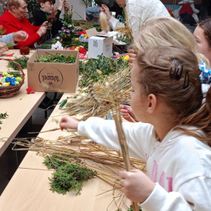 Przy stole siedzą dzieci oraz prowadzące zajęcia, na stole leżą różne materiały potrzebne do wykonania palm wielkanocnych.