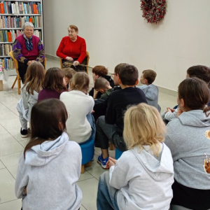 Dzieci siedzą tyłem, przed nimi siedzą prowadzące, jedna z nich czyta książkę.