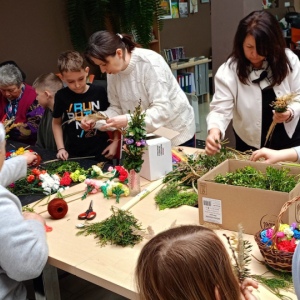 Przy stole stoją dzieci oraz nauczycielki,które pomagają dzieciom w tworzeniu palm wielkanocnych.