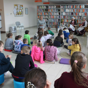 Na dywanikach siedzą dzieci,przed nimi prowadzące siedzą na krzesłąch, jedna z nich czyta książkę.