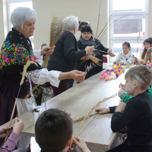 Przy stole siedzą dzieci przed nimi stoją prowadzące, trzymają w rękach materiały potzrebne do wykonania palm wielkanocnych.