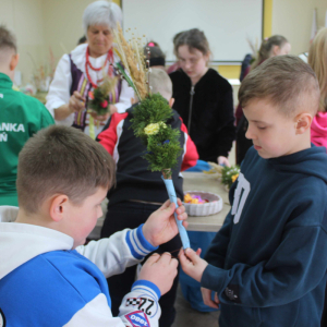 Dzieci oraz prowadzące zajęcia stoją przy stole ,wszyscy robią palmy wielkanocne.