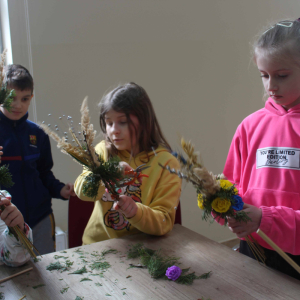 Przy stole stoją dzieci, trzymają w rękach palmy wielkanocne.