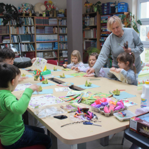 Przy stole siedzą dzieci obok stoi prowadząca, na stole leżą różne kolorowe materiały potrzebne do warsztatów.