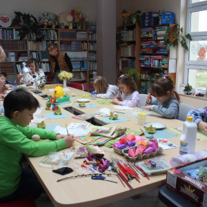 Przy stole siedzą dzieci, obok nich stoi prowadząca, na stole przed nimi leżą różne materiały. Wszyscy malują pisanki.