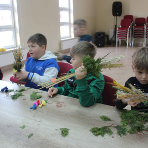 Przy stole siedzi czterech chłopców , trzymają w rękach materiały potrzebne do wykonania palm wielkanocnych.