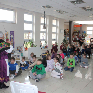 Na dywanikach siedzą dzieci, przed nimi stoi prowadząca zajęcia w ręku trzyma piękną, kolorową palmę wielkanocną.