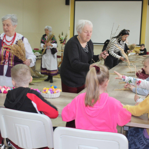 Przy stole siedzą dzieci,przed nimi stoją prowadzące, które pokazują jak wykonać wielkanocne palmy.