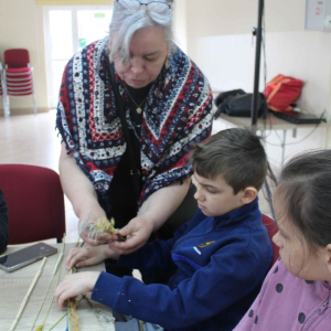 Przy stole siedzi troje dzieci, obok nich stoi prowadząca pokazuje dzieciom jak wykonać palmę wielkanocną.