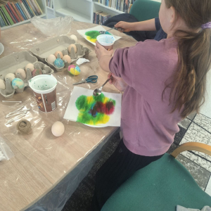 Dziewczynka trzyma w ręku kubek, przed nią na talerzyku leżą papierowe ręczniki pomalowane kolorowymi farbami.