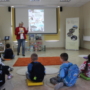 Bibliotekarka trzyma w rękach książkę mówiąc do dzieci siedzących na podłodze