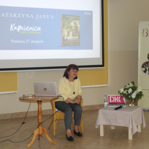 Przy stoliku siedzi pisarka Katarzyna Janus ,za nią stoi duży ekran.