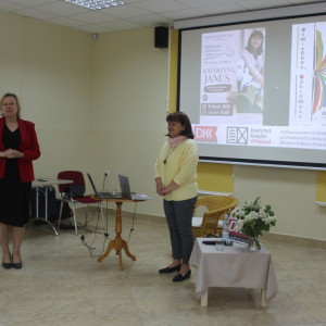 Kierownik biblioteki przemawia do publiczności, obok stoi pisarka Katarzyna Janus. Za nimi wyświetlany jest slajd z plakatami i informacjami dotyczącymi wydarzenia.