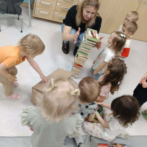 Dzieci wraz z bibliotekarką budują wieżę z książek.