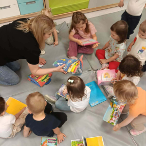 Na materacu siedzą dzieci oraz prowadząca zajęcia wszyscy oglądają kolorowe książki.