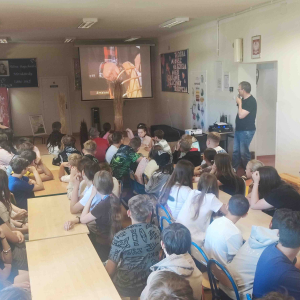 Na dużej sali przy stolikach siedzą uczniowie, obok nich stoi prowadzący zajęcia, wszyscy patrzą na duży ekran przed nimi , na którym wyświetlana jest prezentacja na temat wikliny.
