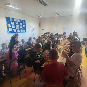 Na dużej sali przy stolikach siedzą uczniowie ,przed nimi na stołach leżą materiały potrzebne do zajęć.