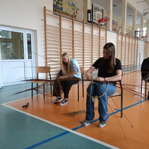Dwie dziewczyny i chłopiec pletą dno do kosza wiklinowego.