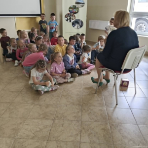 Bibliotekarka siedzi na krześle przed grupą dzieci siedzących na podłodze.