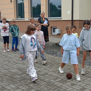  Grupa dzieci idą po chodniku i rzucają kulą z wikliny. Pani prowadząca idzie za nimi. 