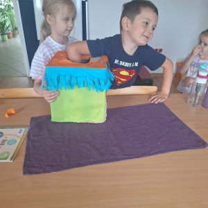 Dziewczynka trzyma kolorowe pudełko stojące na stole,chłopiec wkłada do niego rękę.