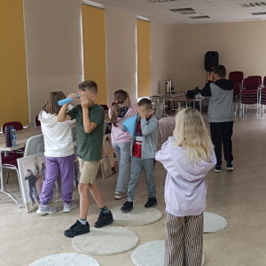 Uczestnicy zajęć chodzą po sali konferencyjnej,jednym okiem patrzą przez tuby wykonane z papieru kolorowego.