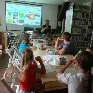 Bibliotekarka tłumaczy dzieciom siedzącym przy stole jak wykonać pracę plastyczną.