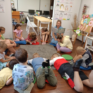Grupa dzieci siedzi w kręgu na podłodze, pani bibliotekarka czyta im książkę.