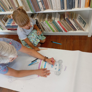Bibliotekarka pomaga dziewczynce rysować jej portret na dużym białym arkuszu